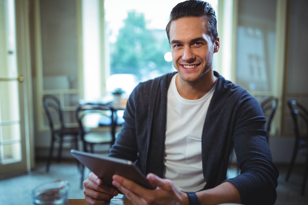 Homem sorridente sentado em um café, segurando um tablet nas mãos, olhando diretamente para a câmera.